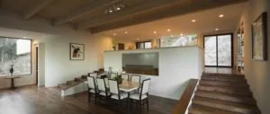 Modern dining area with wooden table and white chairs beneath large fireplace, adjacent to staircase and windows