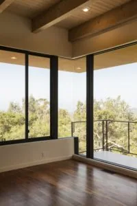 Modern interior room with wooden beams, large black-framed windows overlooking forested landscape, wooden flooring, and metal railing on balcony.