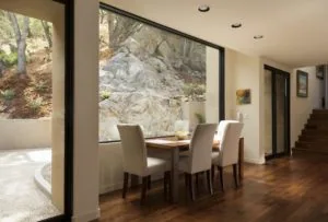 Modern dining area with two upholstered chairs at a table, large windows overlooking rocky hillside, wooden flooring, and recessed ceiling lights.