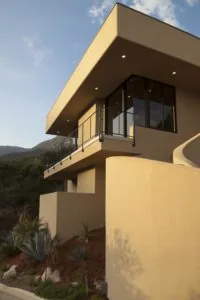 Modern beige stucco residential building with cantilever balcony, large windows, metal railings, and desert landscaping with mountains in background