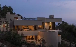 Modern multi-level residential home with curved concrete walls and illuminated interior spaces at dusk, surrounded by native vegetation and hillside landscaping.