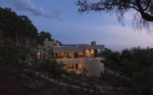 Modern white multi-level house with illuminated interior nestled among trees on a hillside at dusk