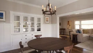 Dining room with round dark wood table, upholstered chairs, white glass-front cabinetry, brass chandelier, and view into adjacent living room