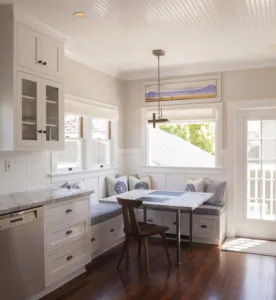 Bright kitchen with white cabinetry, marble countertops, built-in bench seating with cushions and pillows, wooden table, and glass doors opening to outdoor deck.