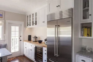Modern kitchen with white cabinetry, stainless steel refrigerator, wood countertop, and glass-front upper cabinets with white beadboard ceiling