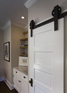 White barn door with black hardware on track next to white cabinetry with marble countertop and wooden shelves