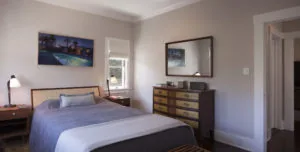Modern bedroom with queen bed, wood dresser with mirror, nightstands with lamps, and framed artwork on walls