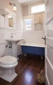 Bright bathroom with white subway tile walls, pedestal sink, white toilet, navy blue clawfoot tub, dark wood flooring, and natural light from two windows