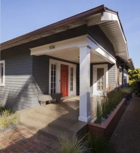 Modern home exterior with gray shingle siding, red front door, white covered porch, and brick entrance steps