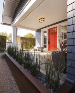 Modern home entrance with red door under covered porch, white shingle siding, and landscaped planter with ornamental grasses