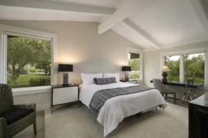 Modern bedroom with white bed, black nightstands, exposed ceiling beams, and large windows overlooking green landscape