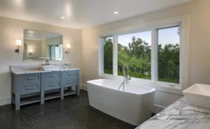Modern bathroom with gray double vanity, large mirror, freestanding white bathtub, and expansive windows overlooking trees