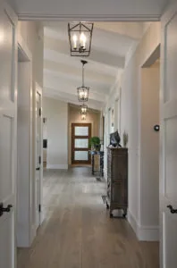 Long hallway with white walls, wood flooring, white paneled doors on either side, and geometric pendant lights hanging from vaulted ceiling.