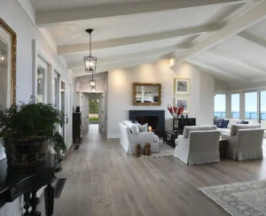 Bright open-concept living room with white shiplap ceiling, exposed beams, fireplace, ocean view windows, white upholstered furniture, and hardwood flooring.