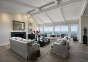 Bright coastal living room with white shiplap ceiling, cream upholstered seating, black fireplace, and expansive windows overlooking ocean views