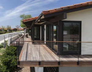 Modern residential deck with wooden flooring, metal cable railings, and large black-framed windows overlooking landscaped grounds under blue sky