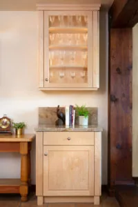 Light wood cabinet with glass door displaying glassware, topped with stone surface holding books, decorative items, and potted plant