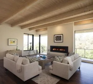 Modern living room with exposed wood beam ceiling, two cream armchairs facing a linear fireplace, large windows with black frames, and hardwood flooring.