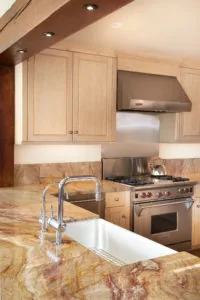 Modern kitchen with light wood cabinetry, granite countertops, stainless steel range and hood, white apron sink with chrome faucet, and recessed lighting.