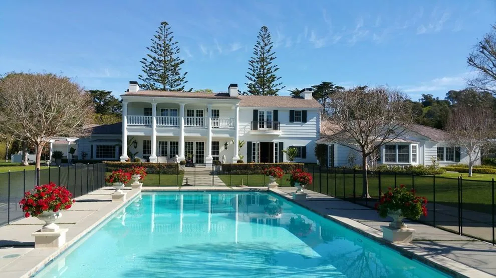 Modern white multi-story house with columned front porch overlooking rectangular swimming pool, manicured lawn, and tall pine trees.