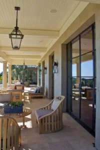 Covered patio with wooden furniture, large black-framed sliding glass doors, pendant lighting, and view of landscaped grounds