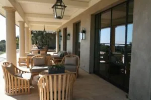 Covered patio with wooden furniture, ceiling fans, large black-framed windows, and stone columns overlooking landscaped grounds