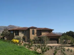 Two-story Mediterranean-style home with tan and brown facade, clay tile roof, arched entryway, attached garage, and mountain backdrop.