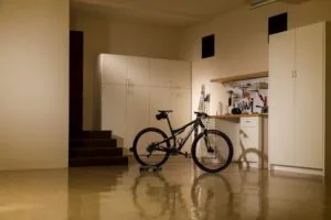 Black mountain bike in modern garage with white storage cabinets, wooden workbench with tools, and polished concrete floor