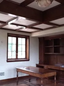 Home office with dark wood coffered ceiling, recessed lighting, wooden desk, built-in shelving, and large window with brown trim.