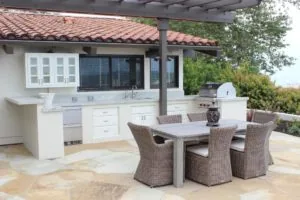 Outdoor patio with white kitchen cabinets, built-in grill, and woven dining chairs around a gray table under pergola