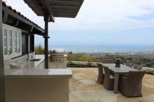 Hilltop patio with outdoor kitchen, dining table, and ocean view overlooking coastal residential area