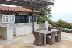 Outdoor patio with white kitchen cabinetry, grill, pergola, dining table and wicker chairs on stone pavers