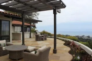 Elevated patio with dark pergola, wicker seating, stone flooring, and hillside views