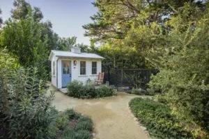 White cottage-style henhouse with blue door and windows, wooden chair, surrounded by landscaping and trees in garden setting.