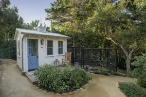 White hen house with blue door and metal roof surrounded by garden landscaping and mature trees