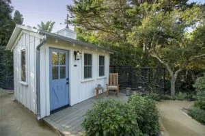 Modern white cottage with blue door, wood deck, and black enclosure surrounded by mature trees and landscaping