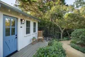 Modern cottage with blue door, deck chair, and black railing surrounded by mature trees and garden plantings