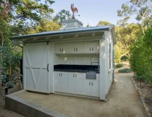 White garden shed with cupola roof topped by rooster weathervane, featuring outdoor kitchen counter with grill, built-in shelving, and barn-style sliding door