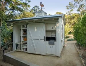 White shed with rooster weathervane, open barn door revealing shelving, and small built-in kitchen area surrounded by trees