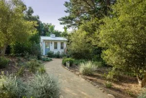 White cottage with blue door nestled among mature trees and native shrubs along a gravel pathway