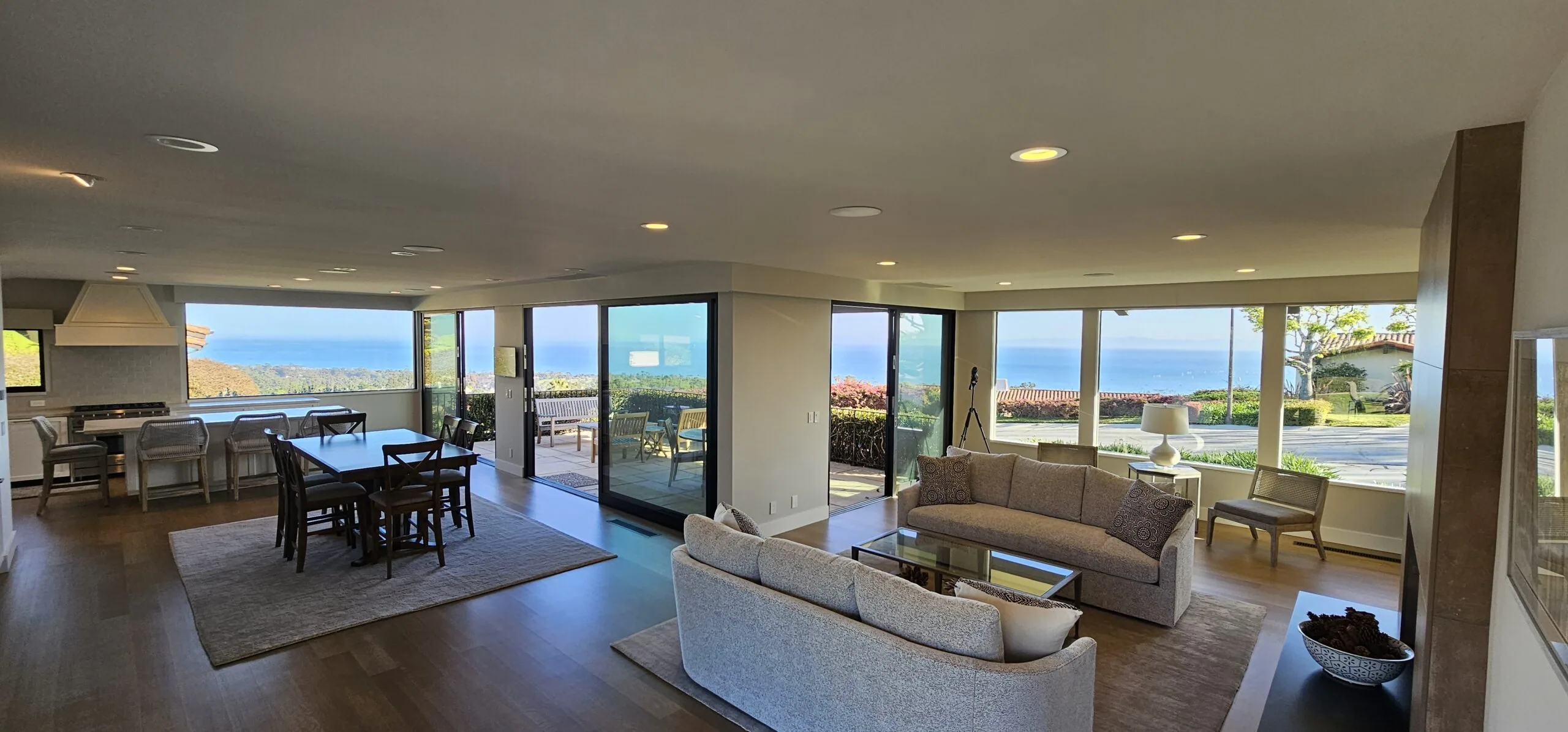 Modern open-concept living space with floor-to-ceiling windows overlooking coastal landscape, featuring dining area, kitchen, and furnished seating