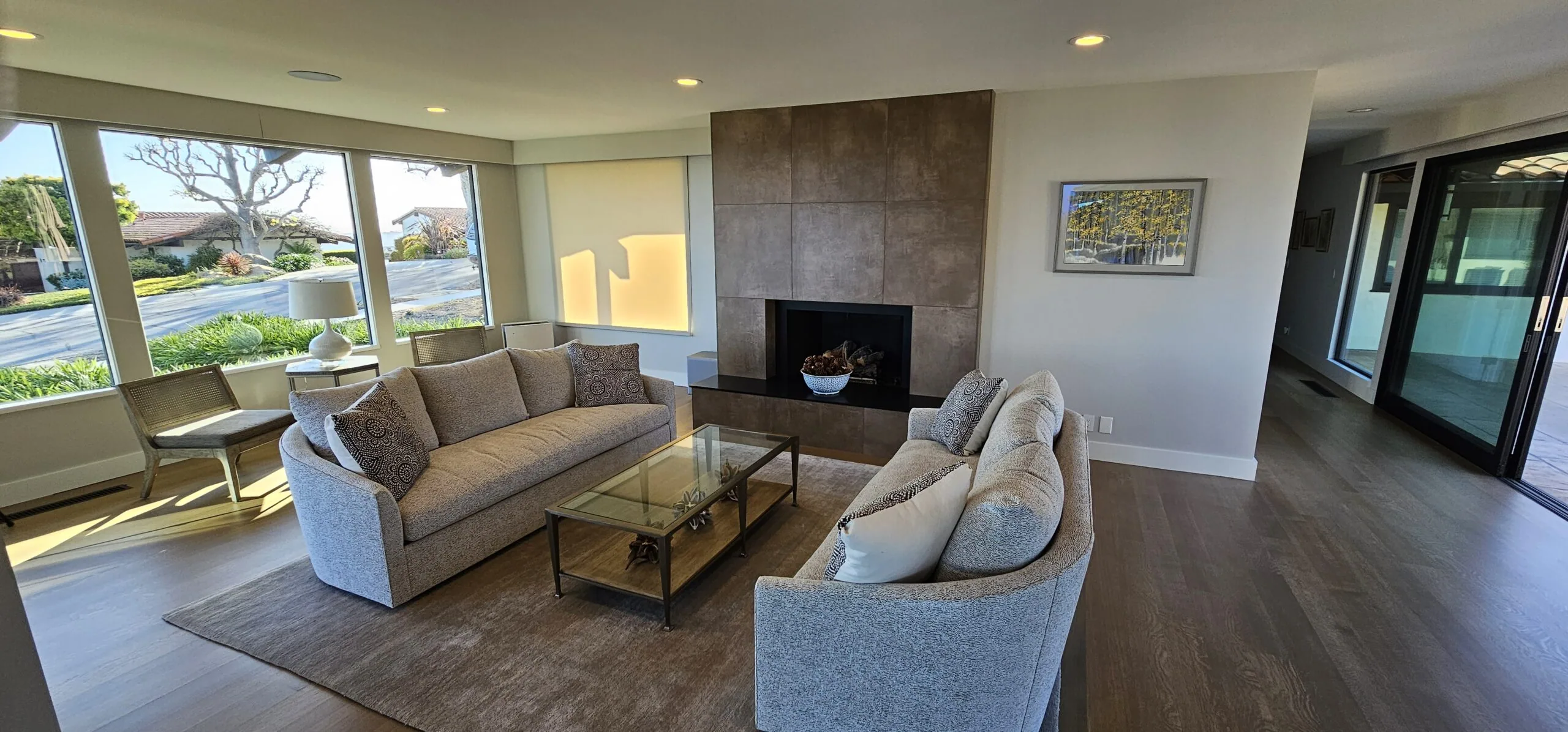 Modern living room with two upholstered sofas facing a fireplace, coffee table, large windows overlooking landscaped exterior, and dark wood flooring.