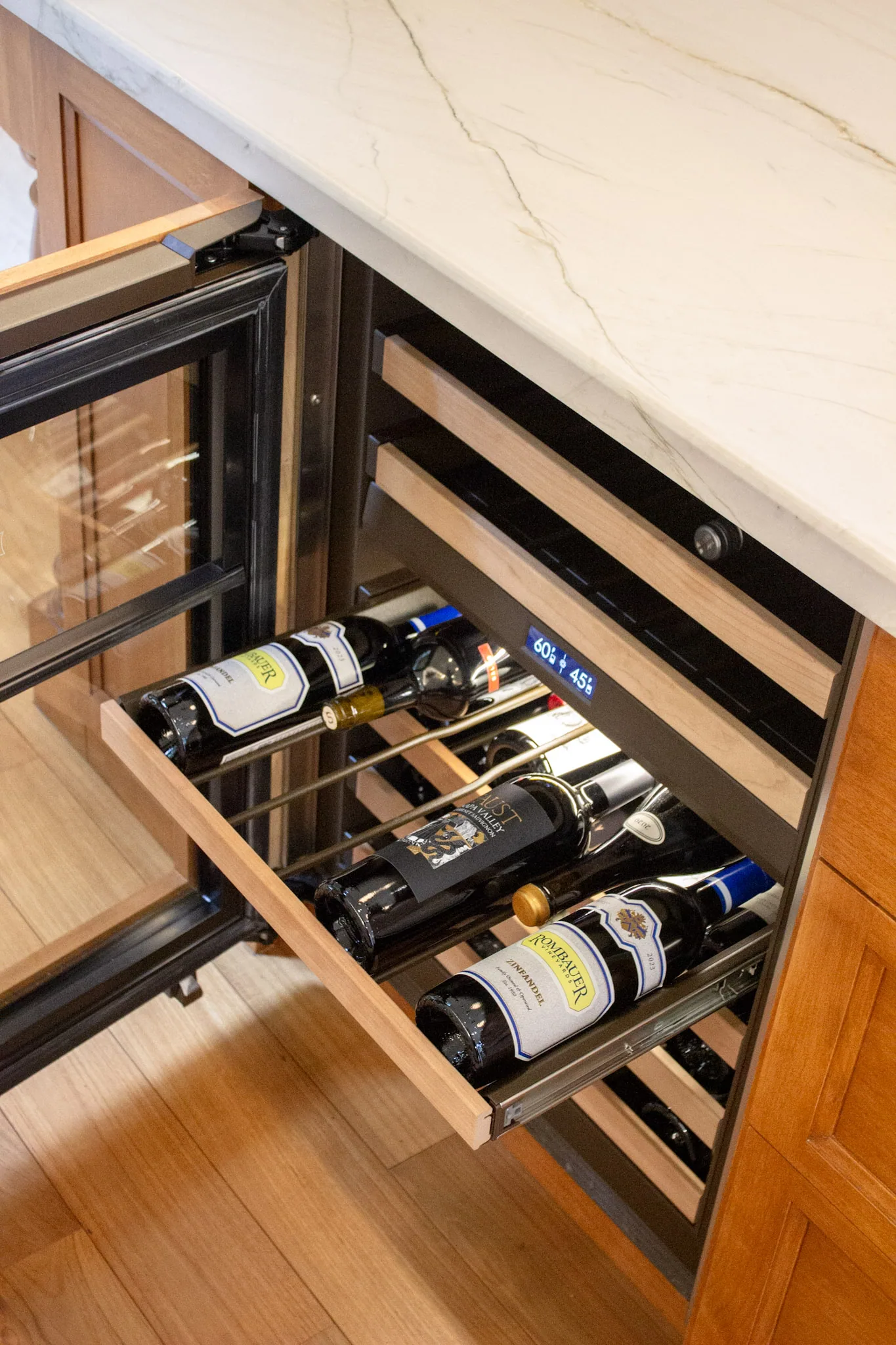 Open wine cooler drawer built into kitchen cabinetry displaying several wine bottles horizontally under a cream marble countertop