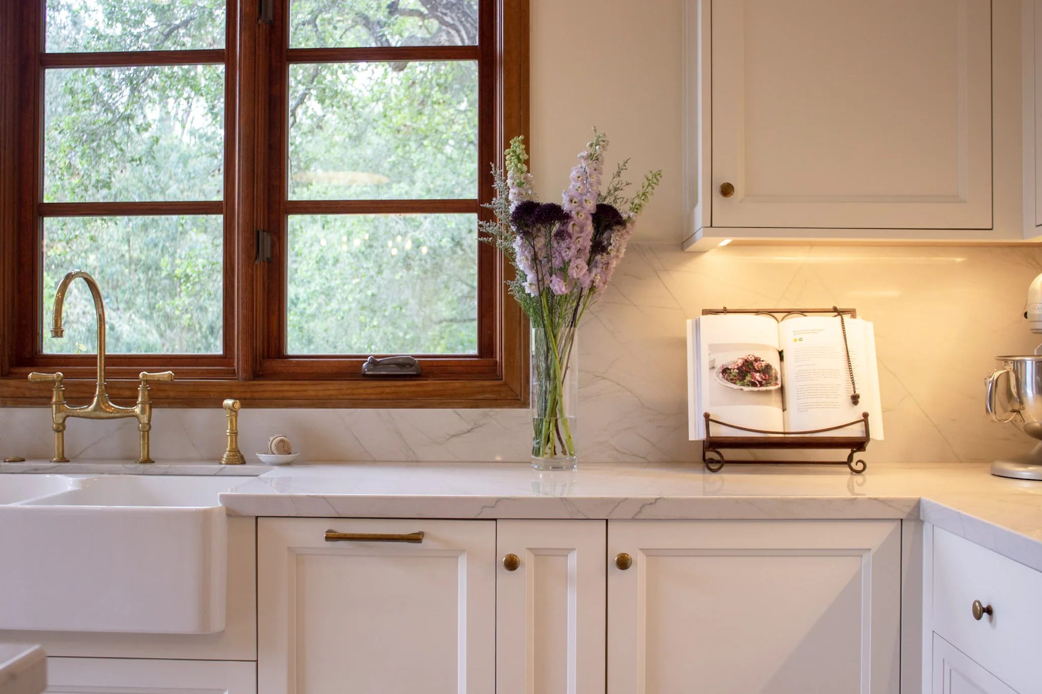 Kitchen sink with white cabinetry, marble countertop, brass fixtures, and dark wood-framed windows overlooking greenery