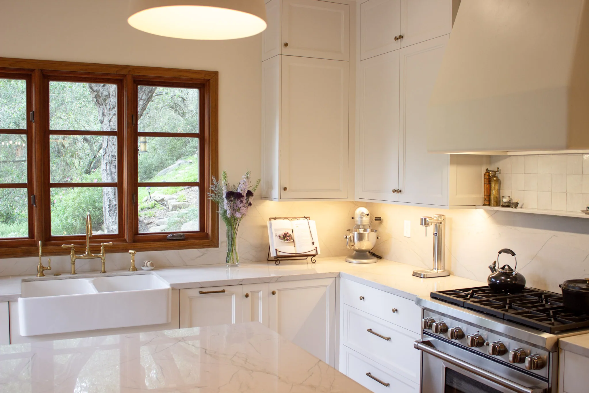 Modern kitchen with white cabinetry, marble countertops, gas range, farm sink, wood-framed windows, and pendant lighting.