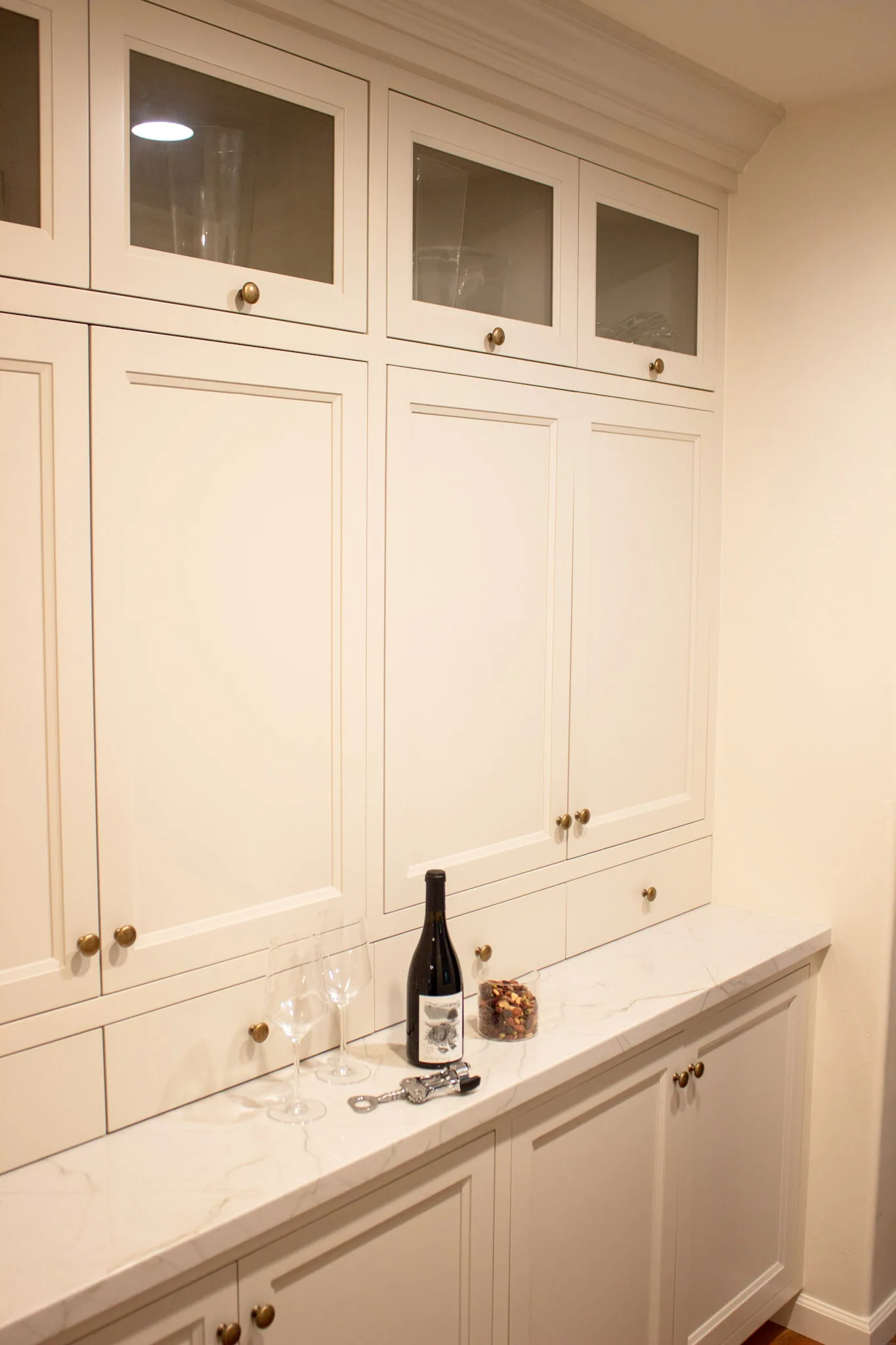 Cream-colored built-in cabinetry with glass-front upper doors, solid lower doors with brass hardware, and white marble countertop displaying a wine bottle and glasses