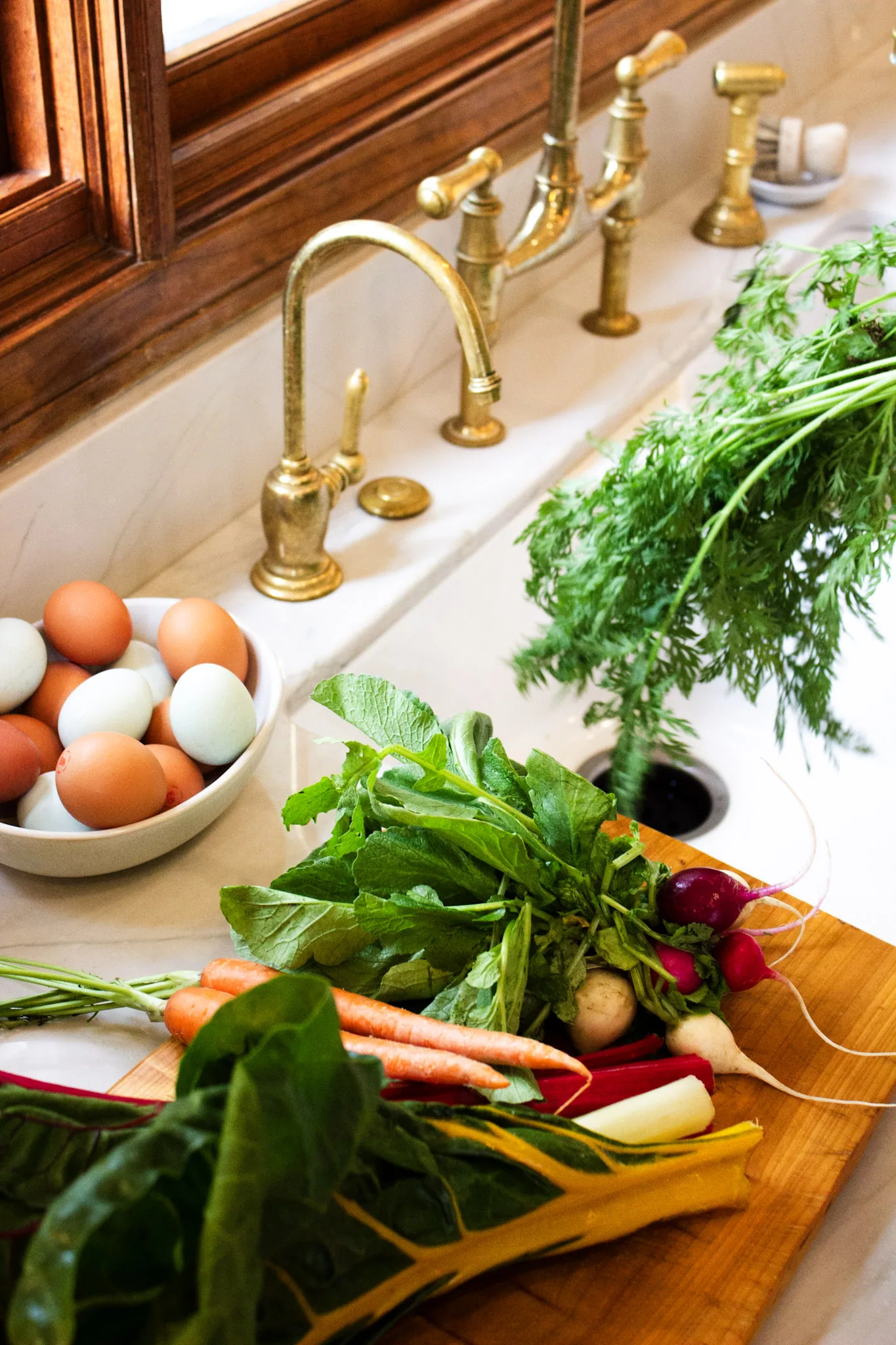 Kitchen sink with brass faucets, bowl of eggs, fresh vegetables on wooden cutting board, and potted herbs