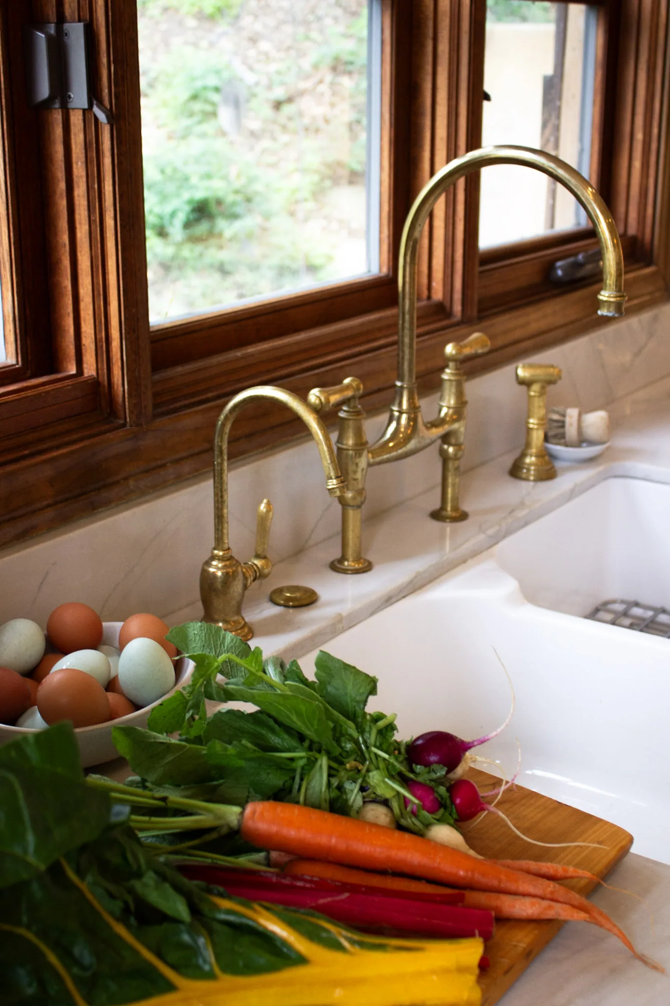Kitchen sink with brass faucets and fresh vegetables on cutting board near window