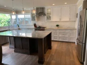 Modern kitchen with white cabinetry, dark wood island, stainless steel appliances, pendant lights, and stone countertops overlooking a backyard.