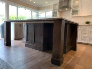 Large kitchen island with dark wood base and built-in dishwasher, cream cabinetry and marble backsplash visible, bright windows overlooking yard